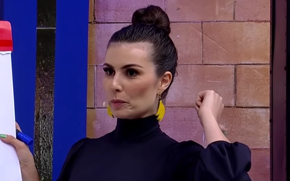 Nathalia Arcuri com um coque no cabelo blusa de gola alta e com a mo indicando para suas costas